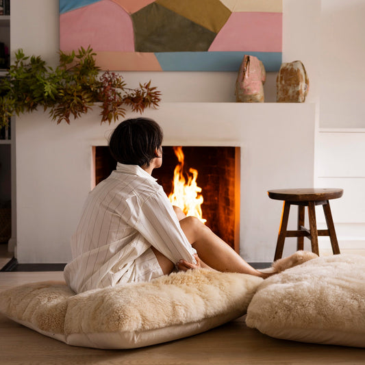 Person sitting on a fluffy alpaca floor pillow in front of a fireplace with abstract art above.