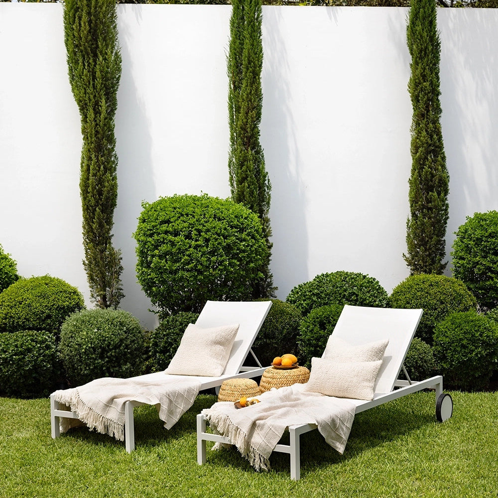 Two white lounge chairs on a grassy area with a white wall and greenery in the background.