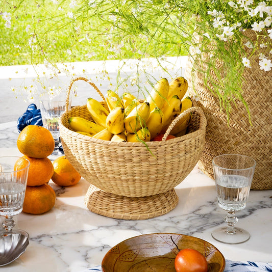 Intiearth Chicha Pedestal Bowl basket full of bananas on a table with flowers and glasses in the background
