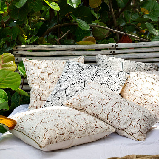 A set of decorative throw pillows with beige and brown organic line patterns arranged on a white daybed outdoors, with a bamboo headboard and lush green foliage in the background.