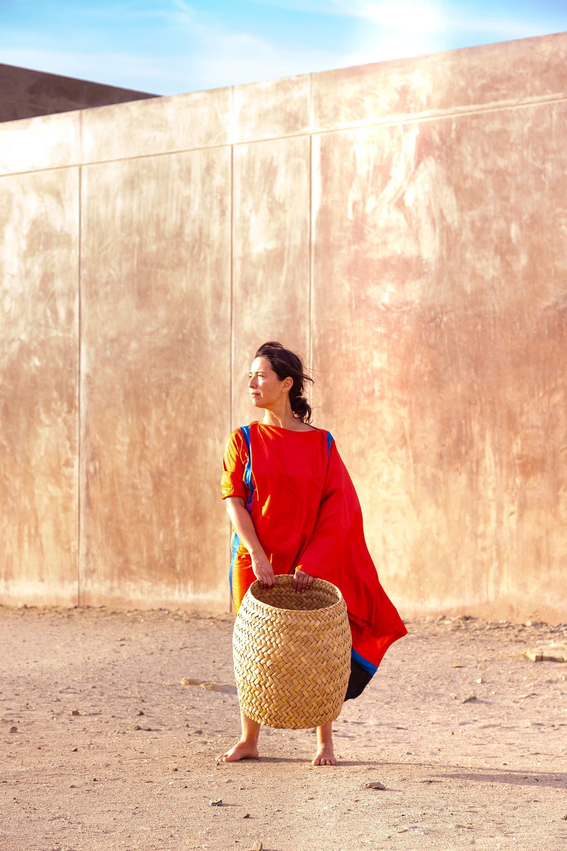 Woman in a red dress holding a woven basket against a beige wall.