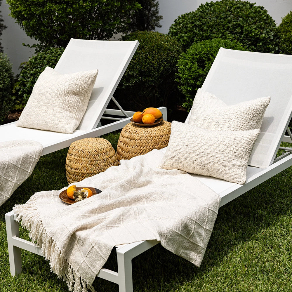 Two white lounge chairs with beige pillows and blankets on a grassy area with greenery in the background.