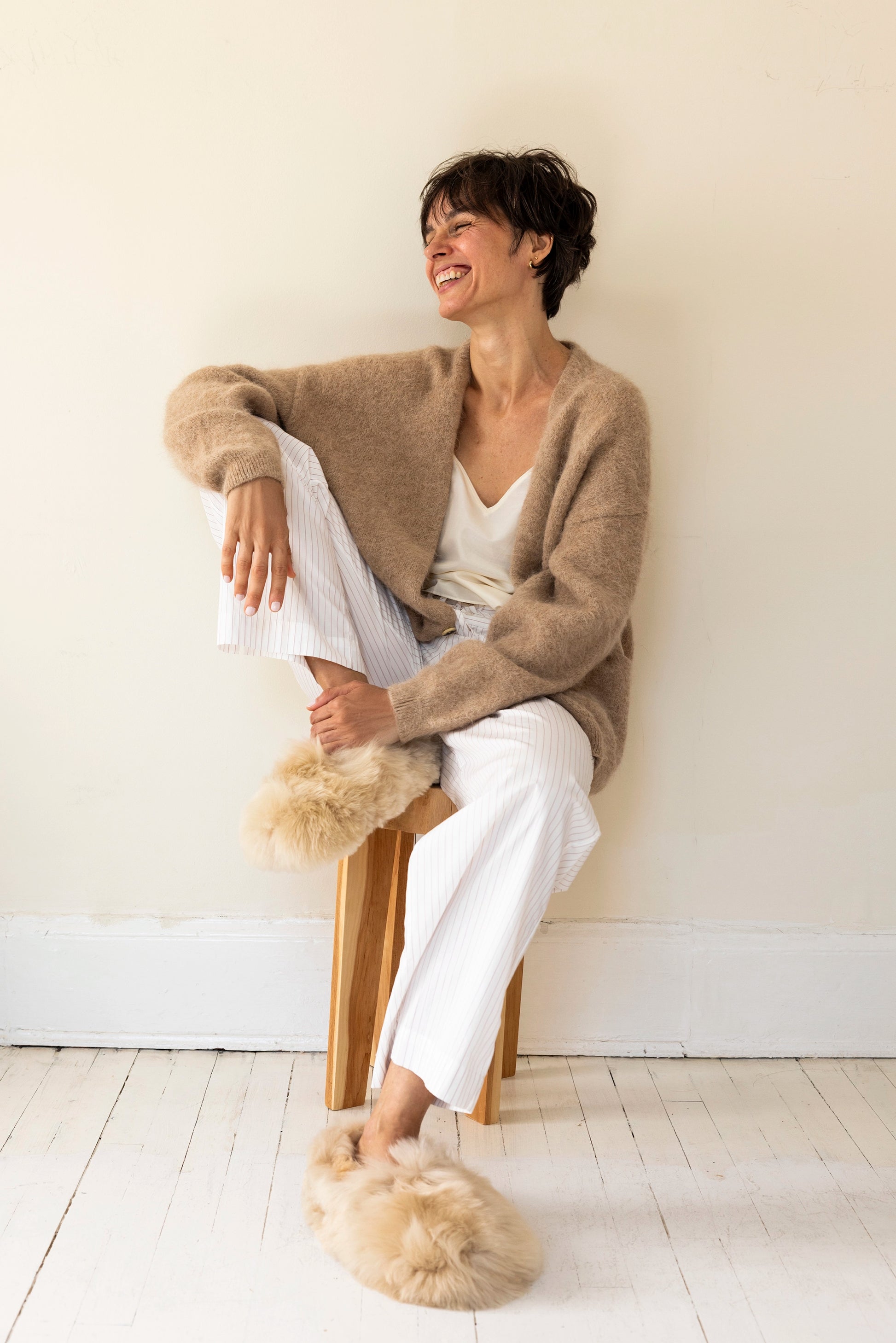 Intiearth Champagne-colored alpaca fur slippers featuring plush fur uppers, soft suede soles, and warm sheepskin insoles on woman sitting on stool