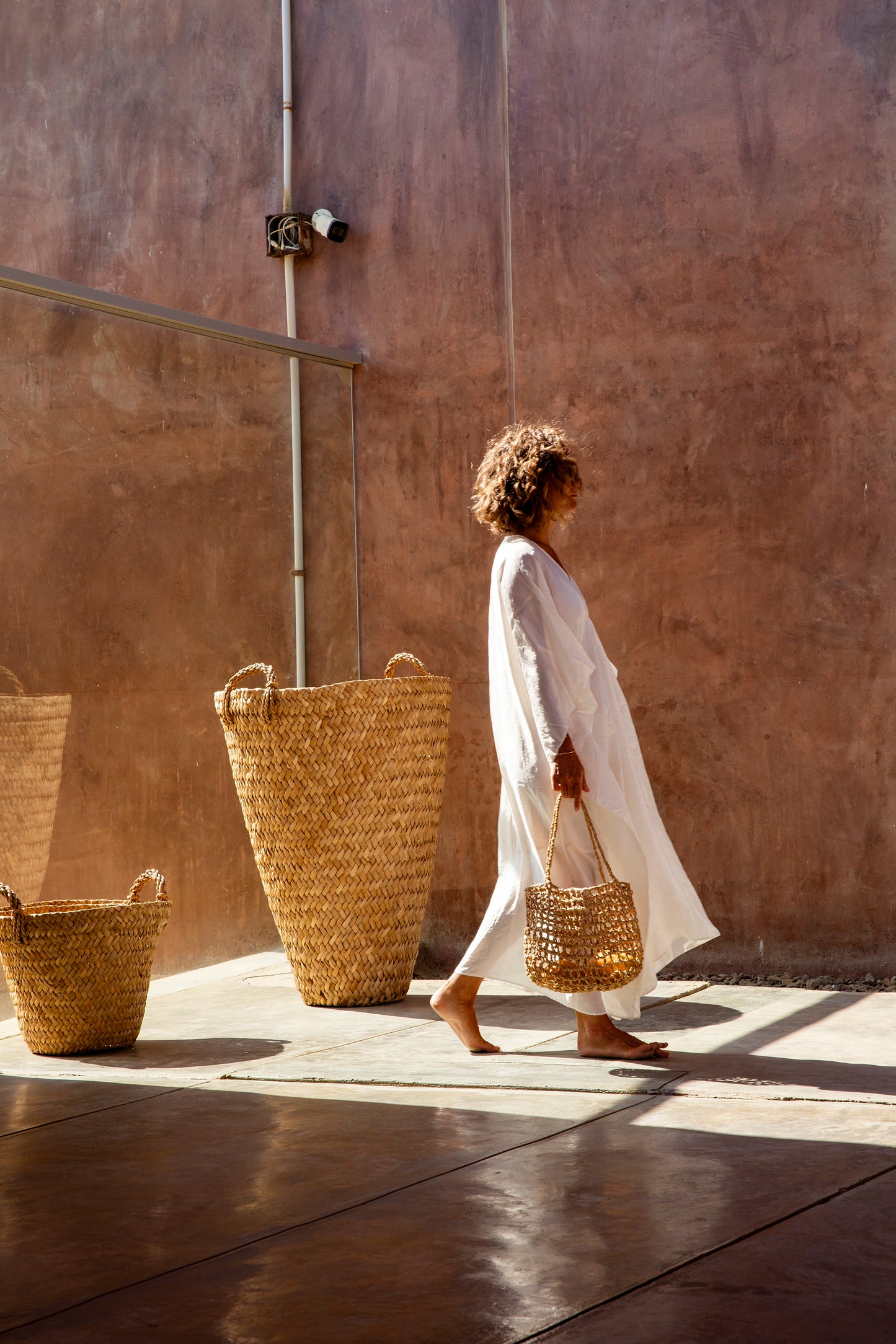 Woman in a white dress holding a woven basket against a brown wall.