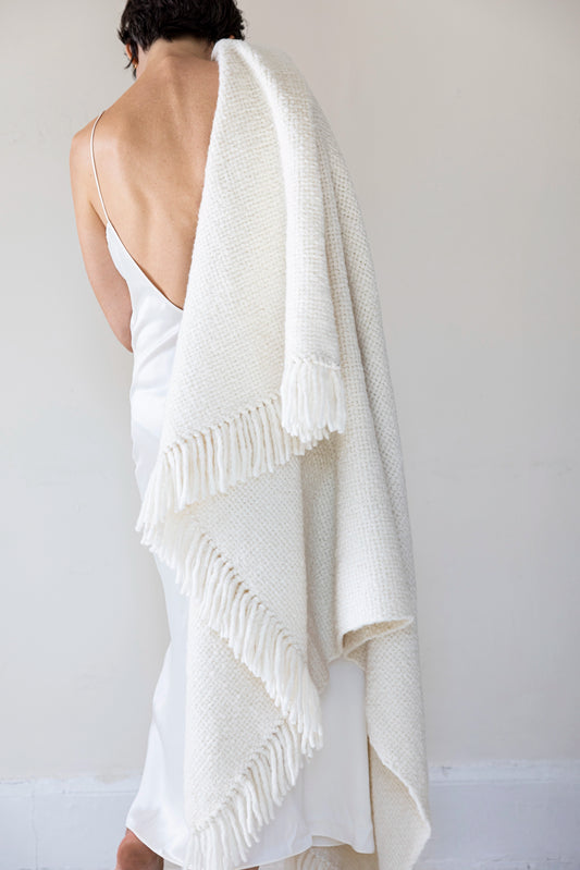 Woman wearing a white dress with a fringed Intiearth hand loomed alpaca throw blanket in ecru draped over her shoulder against a plain background