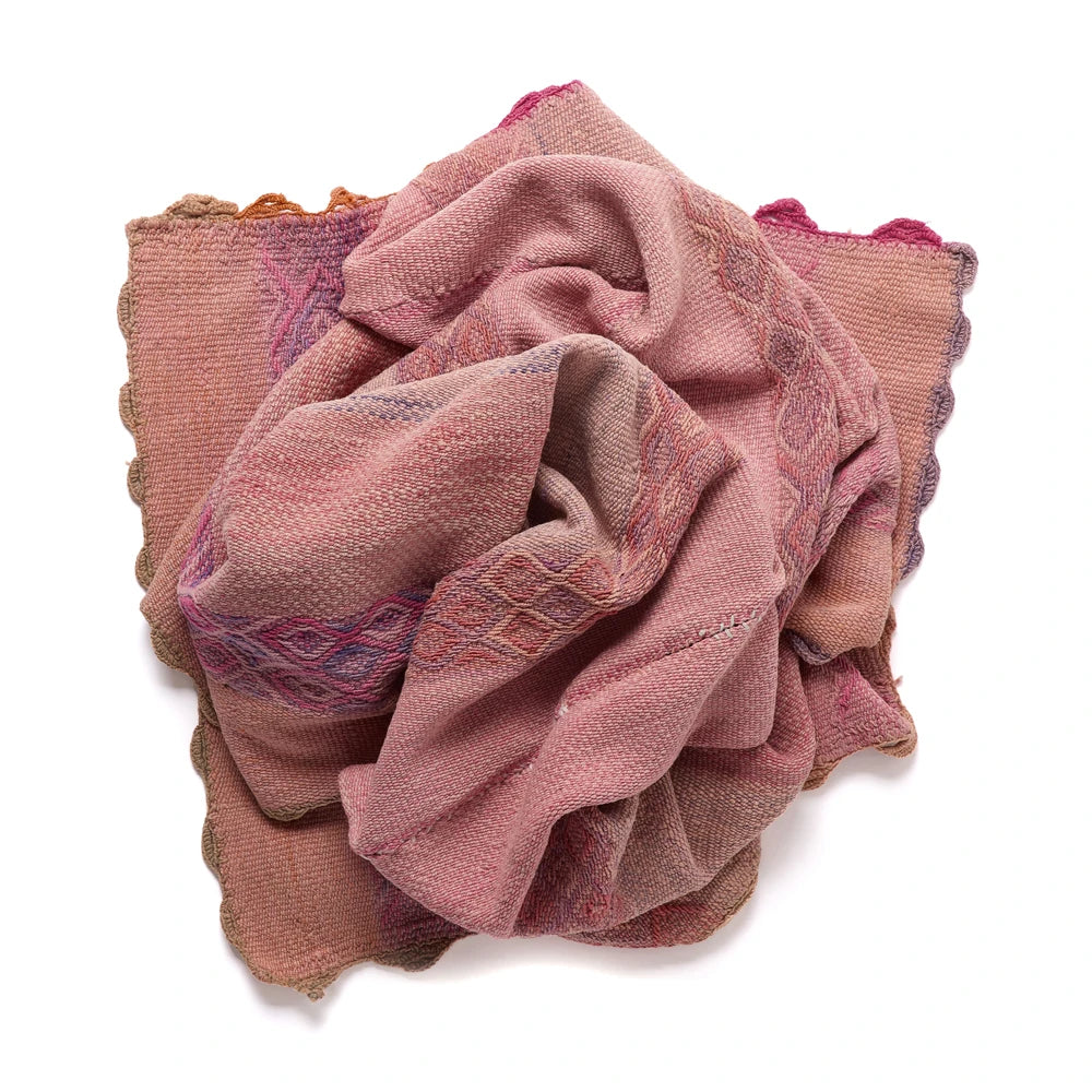A folded woven textile scarf or shawl in dusty pink and mauve tones with purple and blue geometric patterns and scalloped edges, photographed on a white background.