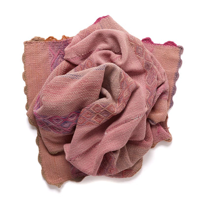 A folded woven textile scarf or shawl in dusty pink and mauve tones with purple and blue geometric patterns and scalloped edges, photographed on a white background.