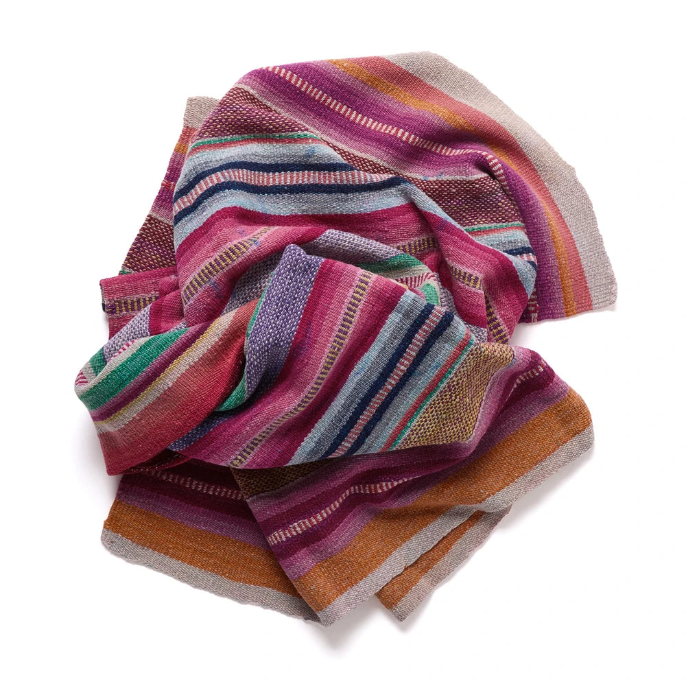A multicolor striped woven textile, likely a throw blanket or scarf, featuring horizontal bands of pink, purple, orange, green, and blue on a white background.