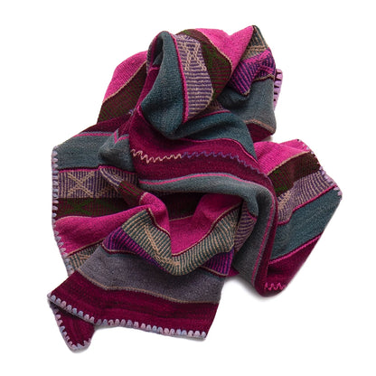 A folded woven textile with horizontal stripes in magenta pink, dark teal, brown, and gray, featuring geometric patterns and a pom-pom trim, shown on a plain white background.