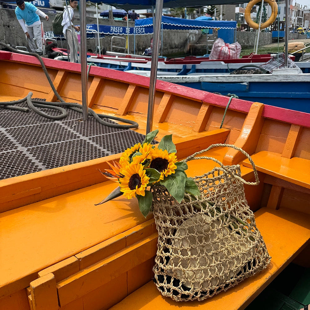 Sunflowers in a woven Intiearth Playa bag on a wooden boat deck with people and boats in the background.