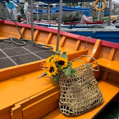 Sunflowers in a woven Intiearth Playa bag on a wooden boat deck with people and boats in the background.