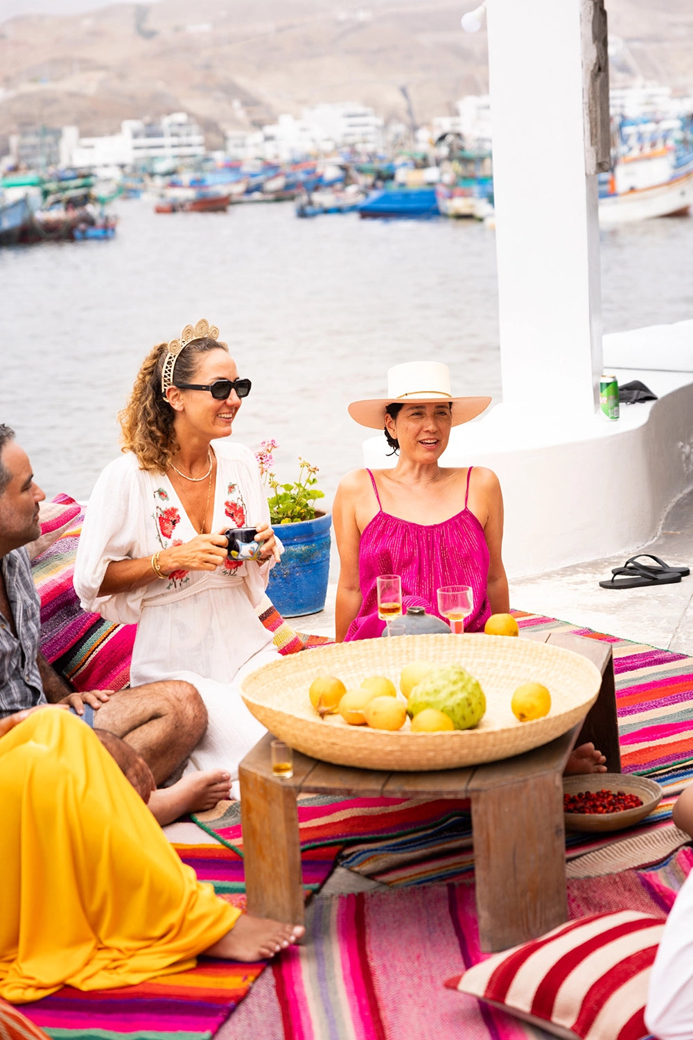 People sitting around a table with fruit on a colorful rug by a waterfront.
