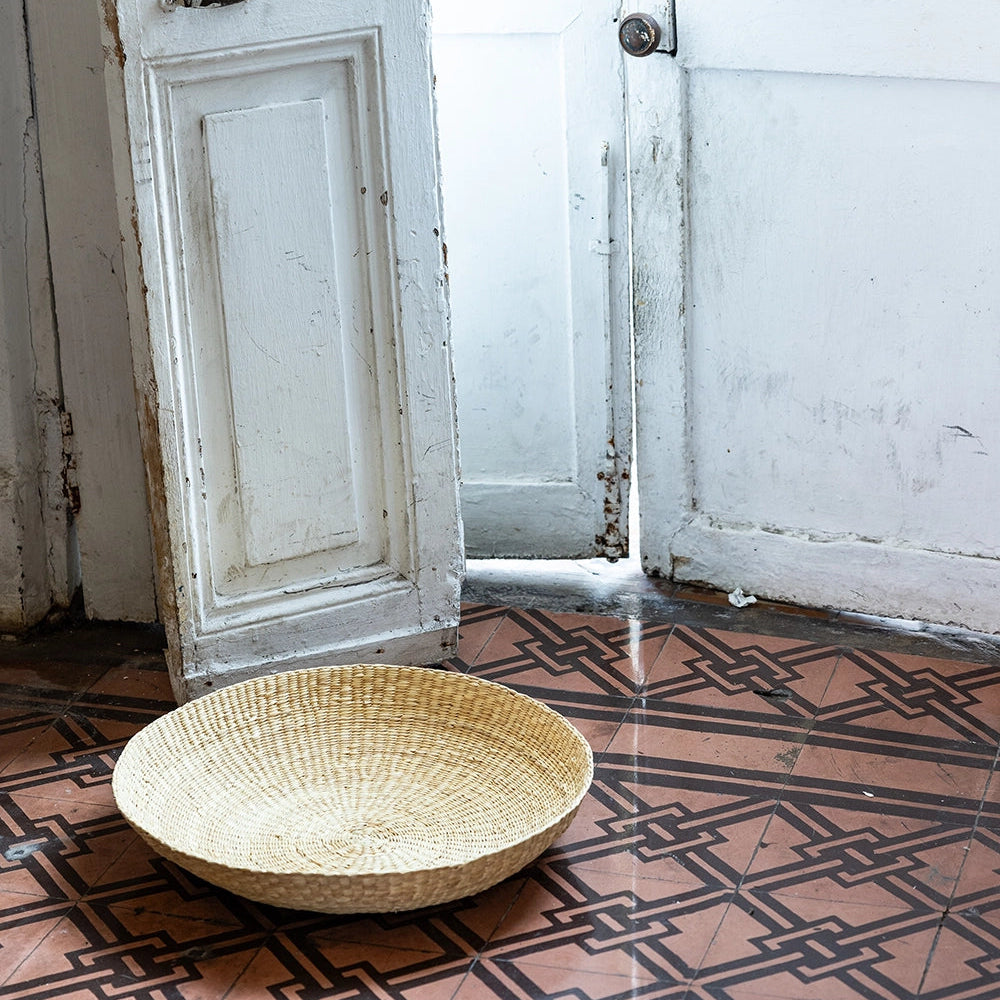 Intiearth El Sol Platter Chica Woven basket on a painted cement tile floor with a white door in the background